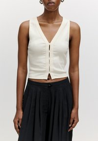 Off-white ärmelloses Oberteil mit tiefem V-Ausschnitt, das eine Vorderknopfleiste und einen verkürzten Saum hat. Weicher Stoff mit leichtem Stretch.