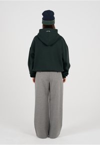 Mørkegrønn oversized hettegenser med hette og ribbestrikkede mansjetter, kombinert med brede grå bukser. Luen er marineblå med grønn stripe.