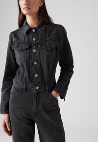 Juodas džinsinis švarkas su užsegimu mygtukais, dviem krūtinės kišenėmis ir prigludusia juosmens dalimi. Oda su smailiu apykakle ir ilgomis rankovėmis.