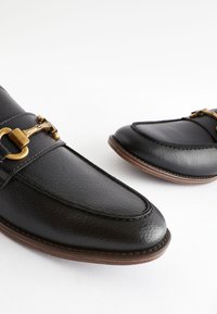 Mocassins en cuir noir avec boucle dorée, texture lisse, bout arrondi et semelle marron avec un détail de couture sur le bord.