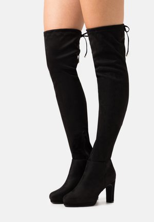 Botas altas negras de ante que llegan por encima de la rodilla, con tacones de bloque, cremalleras en los lados internos y lazos ajustables en la parte superior, usadas con las piernas desnudas sobre un fondo blanco.