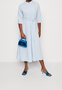 Abito camicia azzurro chiaro con maniche a tre quarti, vita legata e gonna ampia. Abbinato a una borsa blu lucida e sandali con tacco trasparente.