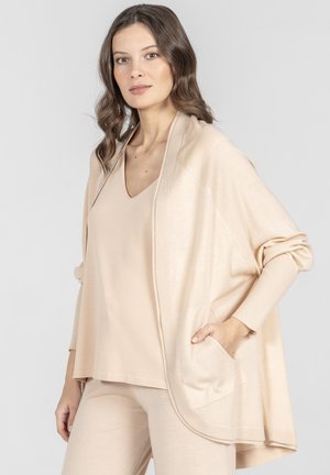 Kobieta ubrana w luźny beżowy kardigan na bezrękawniku i dopasowane spodnie, stojąca z jedną ręką w kieszeni kardiganu na szarym tle.