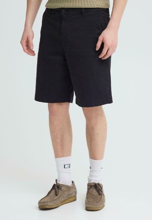 Mand iført sorte knælange shorts, hvide crew-sokker med sort logo og brune ruskindssnøresko, stående på hvid baggrund.