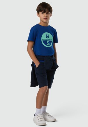 Ragazzo in piedi con le mani nelle tasche che indossa una maglietta blu North Sails, pantaloncini blu scuro, felpa blu scuro annodata in vita, calzini bianchi e scarpe da ginnastica bianche.