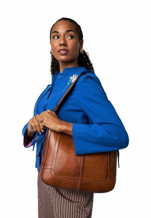 Ruskea nahkainen käsilaukku, jossa on ompelutietoja, kantajalla on sininen takki. Pitkät pystysuuntaiset raidalliset housut ja kukkakirjailu takissa.