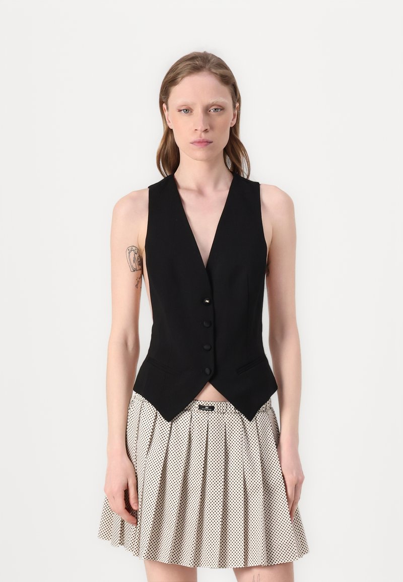 Gilet nero senza maniche con scollatura a V profonda e cinque bottoni, abbinato a una gonna bianca plissettata con pois neri.