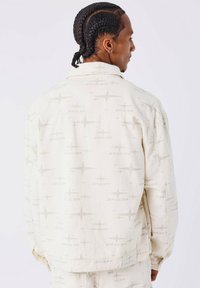 Chaqueta crema hecha de tela con un sutil patrón abstracto. Presenta un corte relajado y un cuello; etiqueta del logo en el dobladillo inferior.