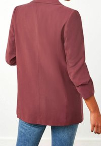 Blaque maroon dengan bahan yang halus, desain bahu yang terstruktur, lengan tiga perempat, dan jahitan belakang yang lurus. Dipakai di atas jeans biru.