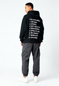 Felpa nera con testo bianco che elenca suggerimenti per il benessere, abbinata a pantaloni cargo grigi e sneakers bianche. Tessuto morbido, vestibilità rilassata, design con cappuccio.