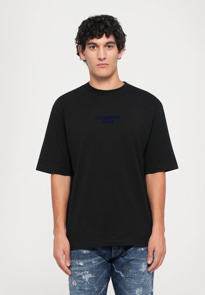 Man with curly hair wearing black DSQUARED2 Milano t-shirt and ripped blue jeans, standing against plain white background.