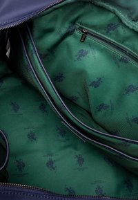 Interiér námořnické látkové tašky má zelenou podšívku s opakujícím se vzorem loga. Obsahuje kapsu na zip a viditelné stehy.