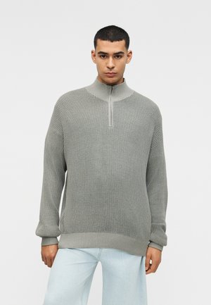 Bărbat tânăr purtând un pulover cu fermoar de jumătate, gri deschis, cu model de nervuri, și blugi albaștri, stând în fața unui fundal simplu, alb.