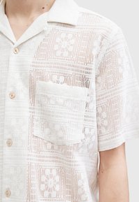 Hvid kortærmet skjorte lavet af et materiale som blonder, med en lomme, knaplukning og et struktureret mønster over hele skjorten.