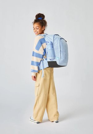 Eine junge Person mit lockigem Haar, das zu einem Dutt gebunden ist, trägt weite beige Hosen, einen blau-creamfarbenen Strickpullover, einen hellblauen Rucksack und passende Sneakers und lächelt seitlich.