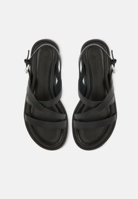 Ζευγάρι μαύρα δερμάτινα σανδάλια με λουριά που διασταυρώνονται και πόρπες στον αστράγαλο, φαίνονται από πάνω σε λευκό φόντο.