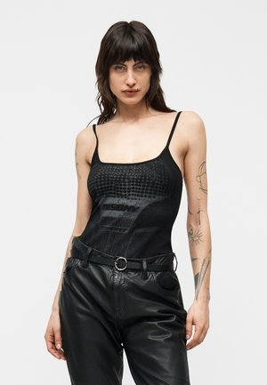 Femme aux cheveux mi-longs foncés et tatouages, portant un haut noir sans manches imprimé et un pantalon en cuir noir avec une ceinture.