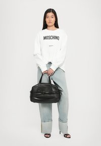 Svart läderhandväska med flera remmar och en slät yta, hålls av en person i en vit Moschino-tröja och ljusblå jeans.