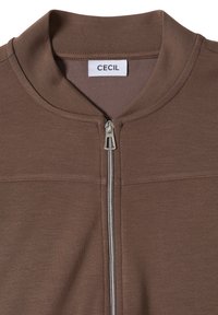 Veste marron à fermeture éclair argentée et col rond, avec une étiquette blanche portant "CECIL" en texte noir à l'intérieur de l'encolure.