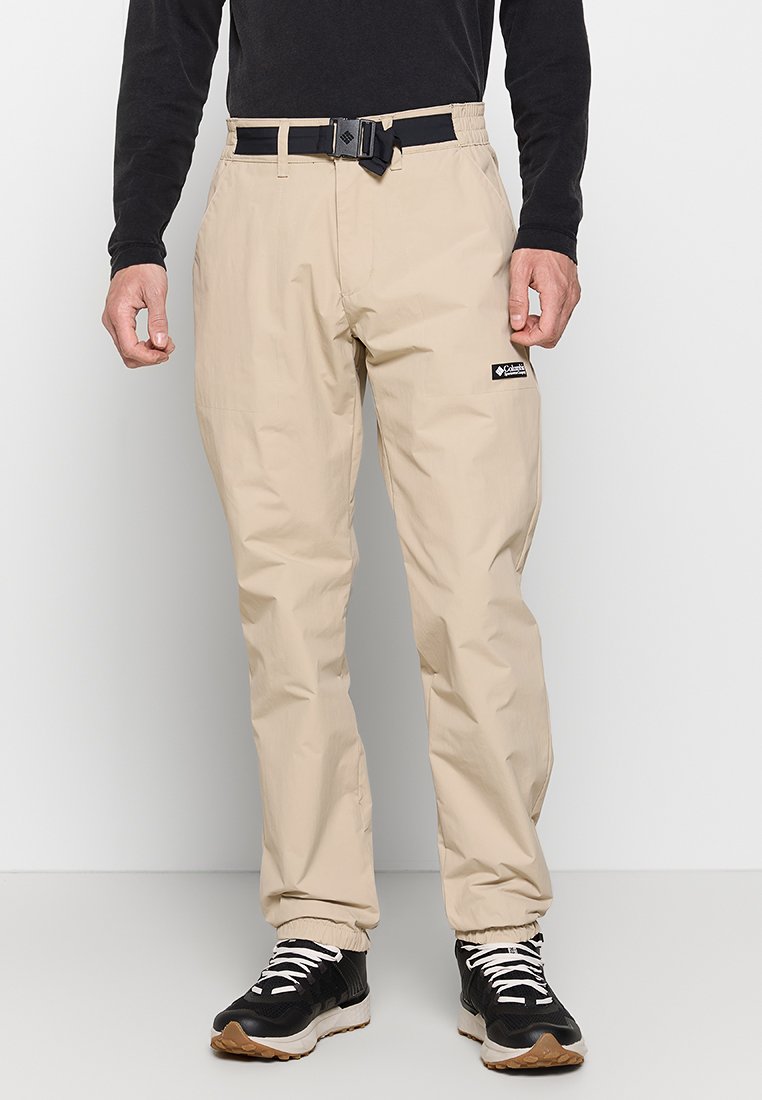 Homme portant un pantalon cargo beige avec une ceinture noire ajustable et des chaussures de sport noires avec des lacets blancs, se tenant contre un mur blanc.