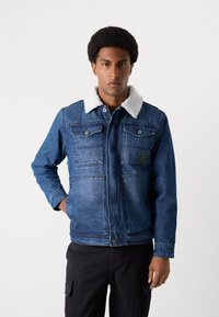Jeansjacke mit einem hellen Schafsfellkragen, zwei Brusttaschen und einem leicht taillierten Design. Dunkelblaue Jeans mit einer strukturierten Oberfläche.