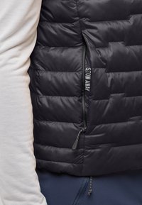 Schwarze Pufferjacke mit gestepptem Design, ausgestattet mit einer seitlichen Reißverschlusstasche und dem Text „STOW AWAY“. Glattes, leichtes Material ist sichtbar.