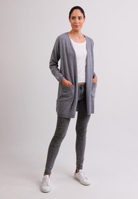 Lange grijze cardigan met verlaagde schouders, open voorkant en voorzakken, gecombineerd met een witte top en grijze spijkerbroek, gedragen met witte sneakers.