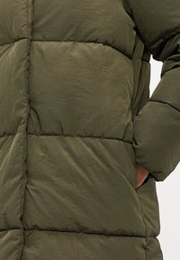Veste matelassée vert olive avec des panneaux cousus, présentant une finition mate et des poches latérales. Texture douce et légère avec une coupe décontractée.