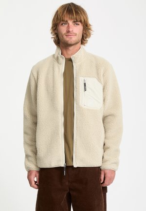 Jeune homme aux cheveux blonds mi-longs porte une veste polaire beige avec une poche poitrine zippée et un pantalon marron.