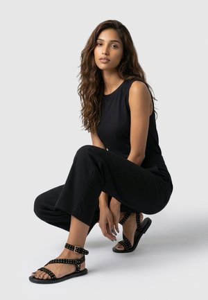 Jonge vrouw met lang, golvend haar, die een mouwloos zwart topje, zwarte wijde broek en zwarte sandalen met studs en enkelbandje draagt, hurkend.