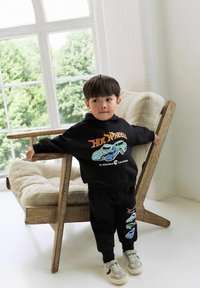 Young boy in black Hot Wheels outfit leaning on wooden cushioned chair near large window with green trees outside.