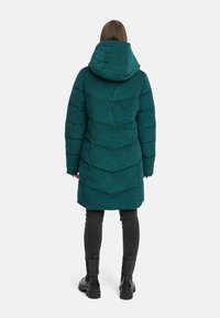 Tealfärgad puffjacka med huva, med en texturerad yta och ett chevronmönster. Buren över svarta byxor och kraftiga svarta stövlar. Bakifrån.