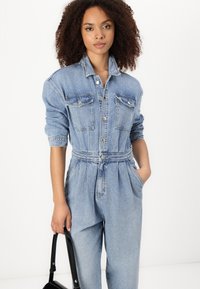 Denim kombinacija v svetlo modri barvi z dolgimi rokavi, z gumbi na sprednji strani, dvema žepoma na prsih in elastičnim pasom. Model drži črno torbico.