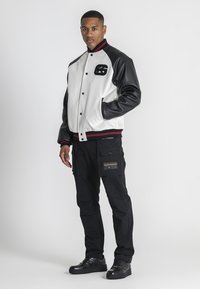Sort og hvid varsity jakke med læderærmer, ribbet krave og et logo. Sammen med sorte cargo bukser og sorte sneakers.
