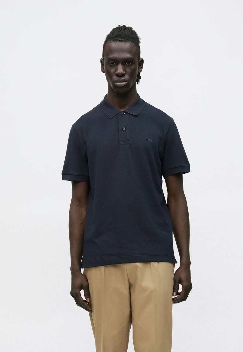 Sötét bőrű férfi tengerészkék pólóingben és bézs nadrágban, egyszínű fehér háttér előtt állva.