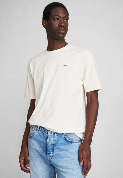T-shirt crème à manches courtes en coton, avec un petit logo noir sur la poitrine. Assorti à un jean en denim bleu clair.
