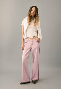 Pantaloni roz deschis cu croială largă și textură fină, asortați cu un top alb scurt, cu detalii de dantelă pe lateral. Încălțămintea este închisă la culoare, cu aplicații decorative.