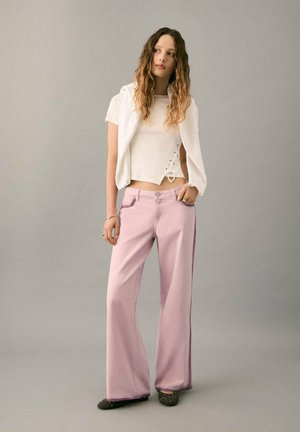 Pantaloni a gamba larga di un rosa chiaro con una texture liscia, abbinati a un top corto bianco con dettagli in pizzo sui lati. Le calzature sono scure con decorazioni.