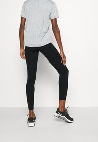 Grå kortärmad skjorta med bakseam, parat med svarta leggings. Modellen bär svarta träningsskor med vita detaljer och synlig dämpning i sulan.