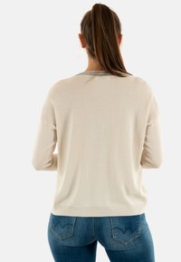 Maglione a maglia beige chiaro con maniche lunghe e scollatura rotonda, con un accento argento attorno al colletto. Indossato con jeans blu.