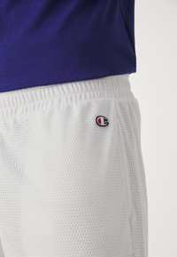 Pantalones cortos atléticos de malla blanca con cintura elástica y un pequeño logo bordado. Llevado con una camiseta morada.