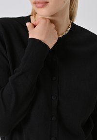 Eine Frau trägt eine schwarze, geknöpfte Strickjacke und eine goldene Kettenhalskette, während sie mit der Hand auf ihrem Kinn über einem schlichten Hintergrund ruht.