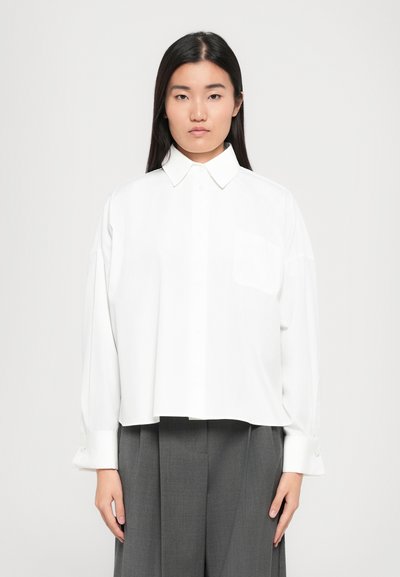 Chemise blanche à col pointu et manches longues, dotée d'une poche poitrine unique et d'un design légèrement cropped.