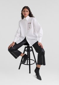Camicia bianca oversize con un design grafico sulla tasca, abbinata a pantaloni cropped in pelle nera con dettagli in zip e stivaletti a pois.