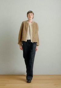 Jachetă maro din blană artificială cu manșete elastice, asortată cu un top alb cu textură reiată și pantaloni închiși la culoare. Ținuta include un fermoar frontal și o siluetă casual.