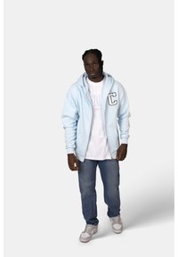 Felpa con zip di colore azzurro chiaro con una grande "C" sul petto, indossata sopra una maglietta bianca, abbinata a jeans di denim e scarpe grigie.