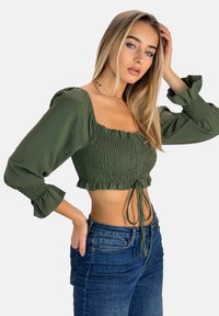 Olivengrøn crop top med sammenkrøllet stof og flæsekant. Lange, puffede ærmer med elastiske manchetter. Lyse jeans, hænderne på hoften.