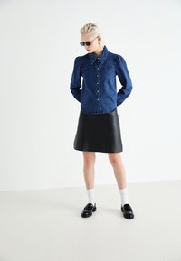 Džinsinė marškinė su pūstomis rankovėmis ir mygtukų uždarymu, derinami su juoda odine mini sijonu, baltais kojiniais ir juodais loafers.