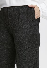 Svarta glittriga byxor med elastisk midja och fickor. Tyget har en texturerad, glänsande yta med silverprickar.