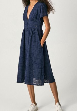 Mujer con un vestido midi de encaje tipo ojo en azul marino, con mangas cortas y bolsillos, combinado con zapatillas blancas, de pie sobre un fondo liso.